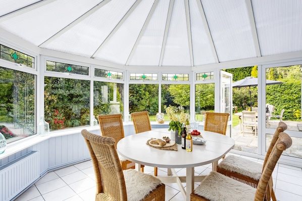 A conservatory with a round table, wicker chairs, and a view of a garden.