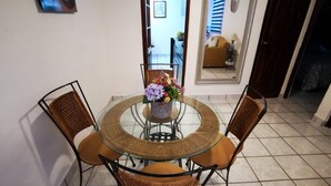 Un coin repas avec une table en verre entourée de chaises en osier, un vase de fleurs et une vue sur un salon avec un canapé et une fenêtre.