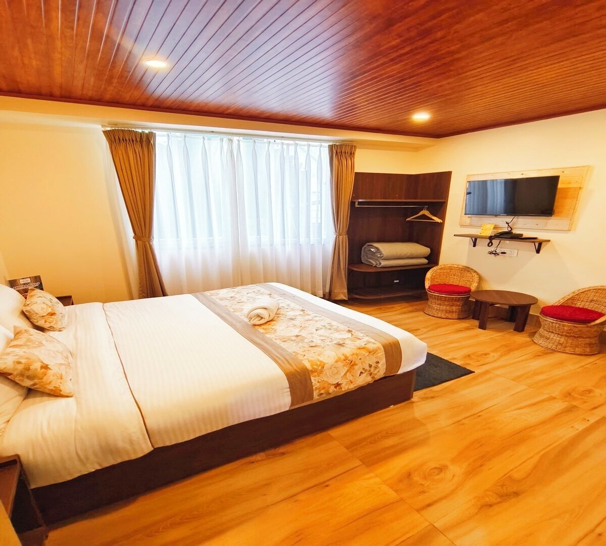 A hotel room with a wooden ceiling, a bed with floral bedding, a TV mounted on the wall, and a seating area with wicker chairs.
