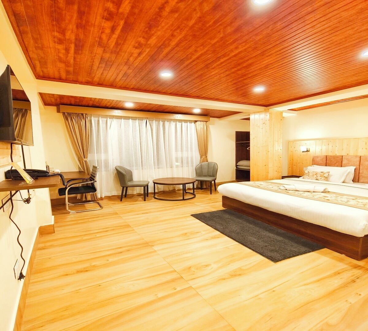 A hotel room with a wooden ceiling, a bed, a desk with a chair, and a small round table with two chairs.
