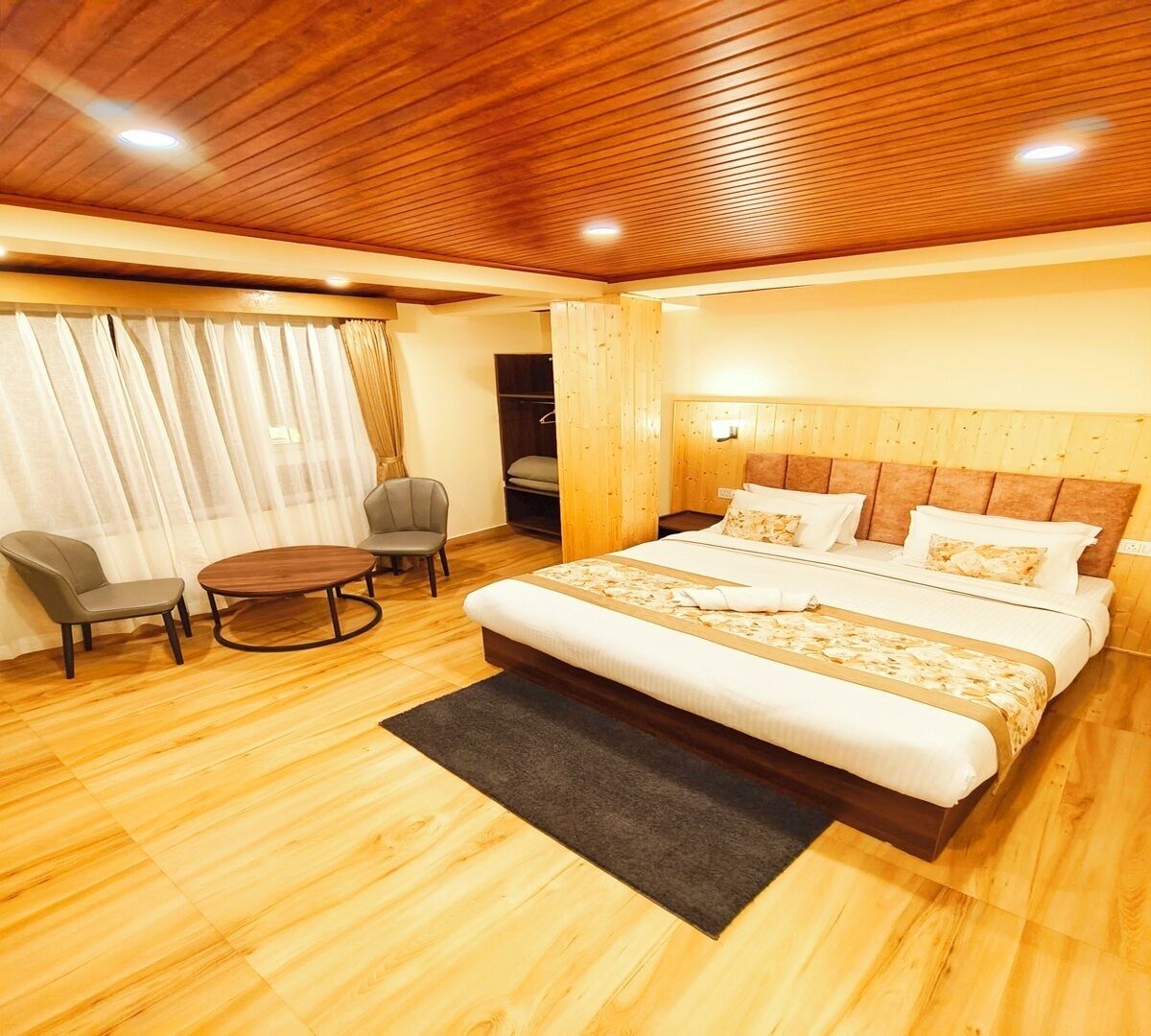 A hotel room with a wooden ceiling, a large bed with two pillows, a small round table, two chairs, and a wooden wardrobe.