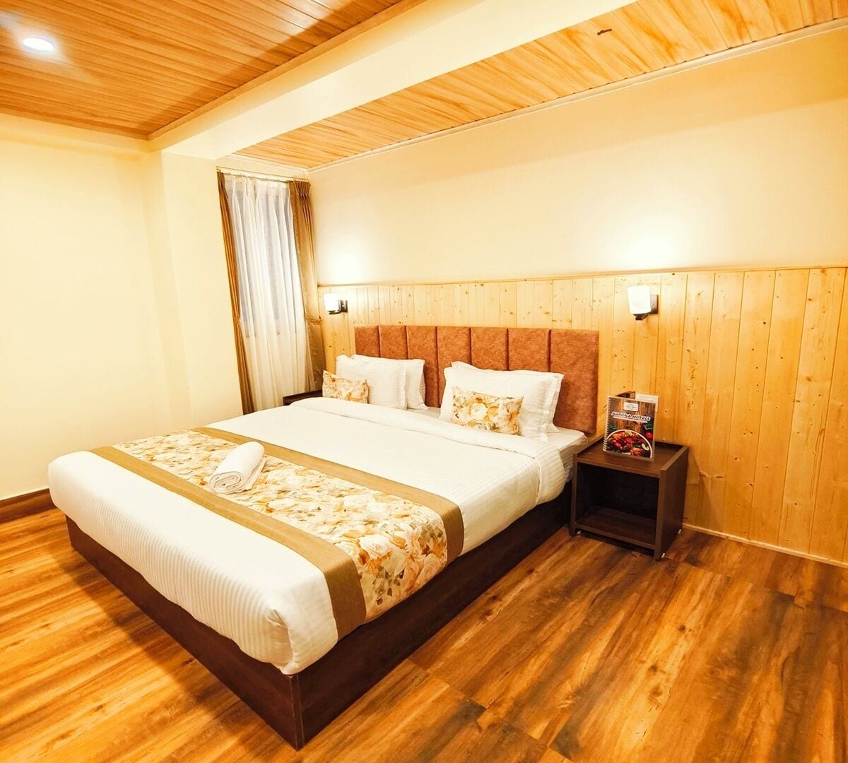 A neatly made bed with a floral bedspread and pillows, a wooden headboard, a nightstand with a magazine, and a wooden floor.