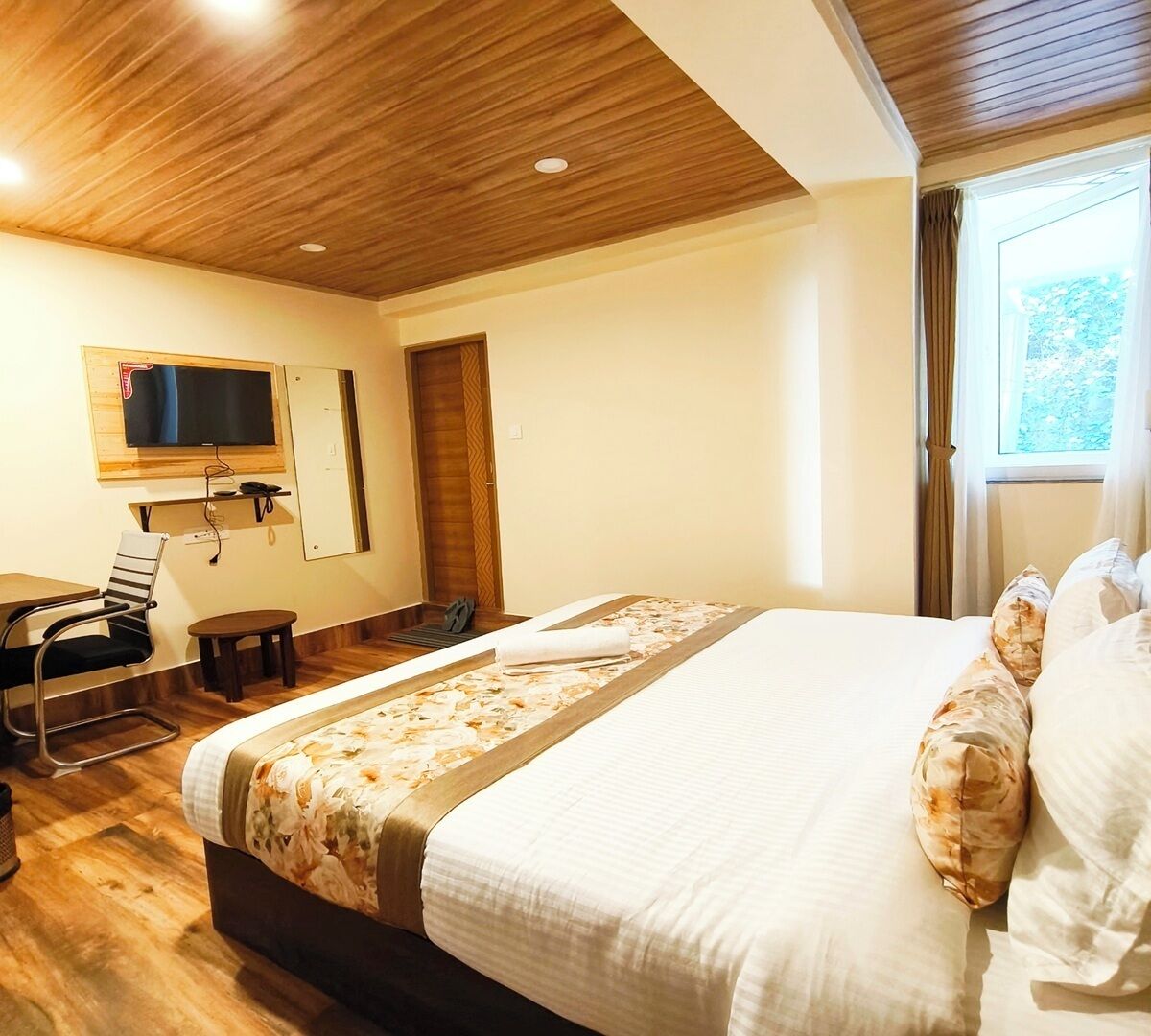 A hotel room with a wooden ceiling, a bed with floral bedding, a TV mounted on the wall, and a window with curtains.