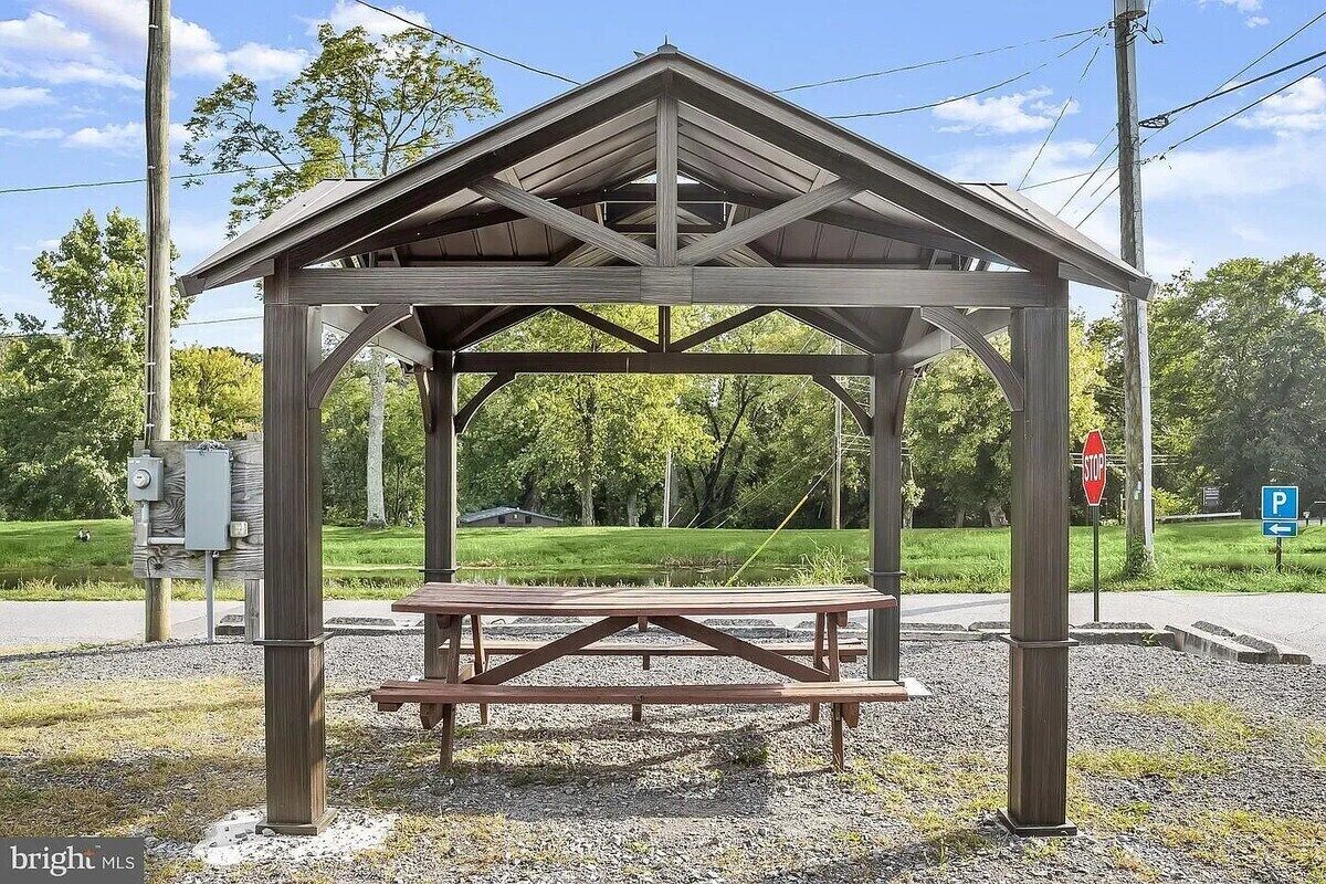 A wooden gazebo with a picnic table in the center, surrounded by gravel and greenery.