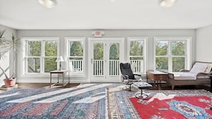Une pièce spacieuse avec de grandes fenêtres, un fauteuil noir, un canapé en osier et un tapis à motifs.
