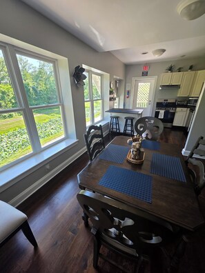 A dining area with a large window, a kitchen in the background, and a view of a garden. - Exclusive House Stay (Hancock)