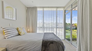 A modern bedroom with a large bed, a bedside table, a framed artwork, and a view of the outdoors through large windows. - Water Views Apartment | Stylish 3BR Villa with Pool & Lake Access (Taupō)