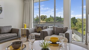 A modern living room with a large window, grey sofas, a small table with a yellow bowl, and a view of greenery. - Water Views Apartment | Stylish 3BR Villa with Pool & Lake Access (Taupō)
