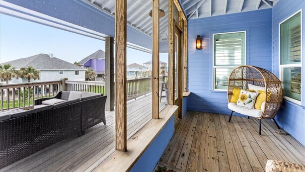 A covered porch with a wicker sofa and a hanging chair, wooden deck, and a view of neighboring houses. - Charming 2-bedroom cabin in serene Crystal Beach (Bolivar Peninsula)