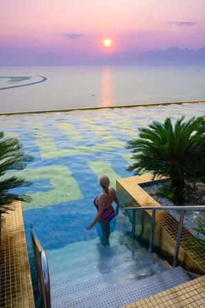A pool with a clear view of the sunset over the water, surrounded by palm trees and a tiled deck. - Apartment hotel at wyndham golden bay có ban công máy giặt (Đà Nẵng)