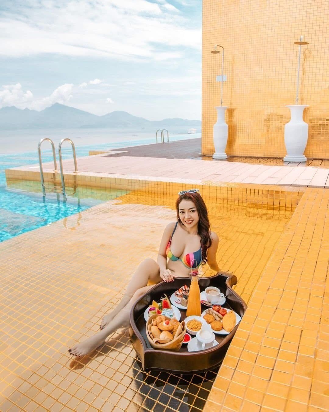 Una dona asseguda al costat d'una piscina amb una bandeja de menjar i begudes, amb vistes a una piscina i a muntanyes al fons.