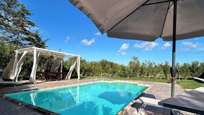 Una piscina con un borde de baldosas, una zona de estar techada y una estructura de pérgola.