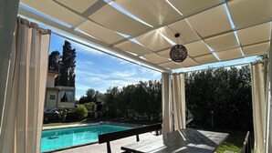 An outdoor patio with a pool, covered seating, and a view of a house and trees. - Villa Eugenie with pool (Sardegna)