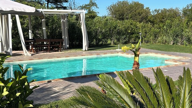 Piscina con toldo blanco y zona de estar, rodeada de exuberante vegetación y un muro de piedra.