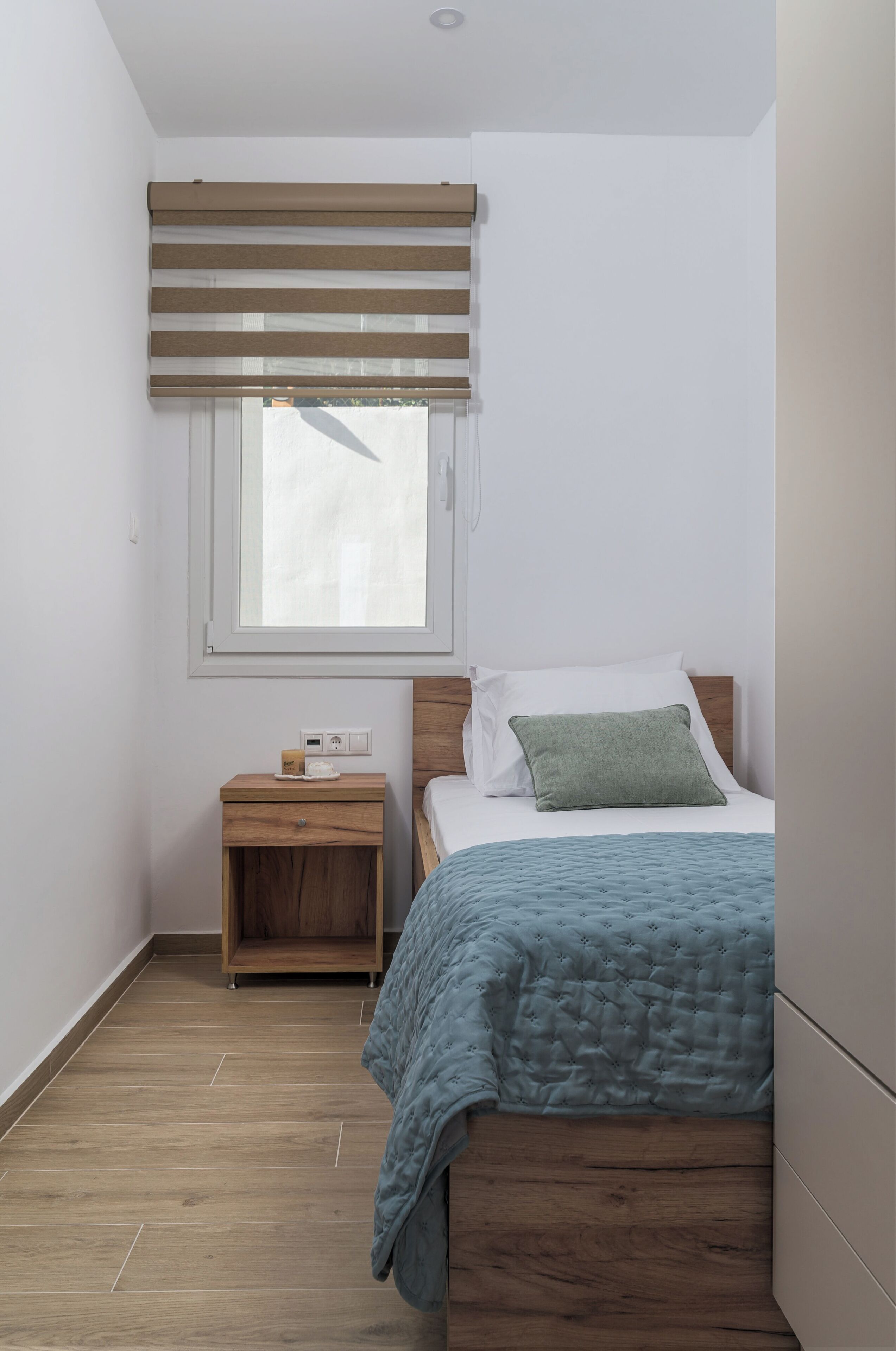 Quarto com uma cama, mesa de cabeceira, janela com persianas e chão de madeira.