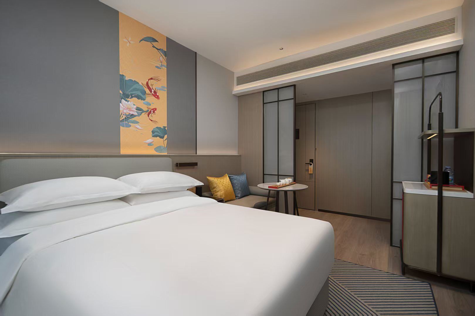 A modern hotel room with a large bed, a small round table with a cup and saucer, a decorative wall piece, and a walk-in closet.