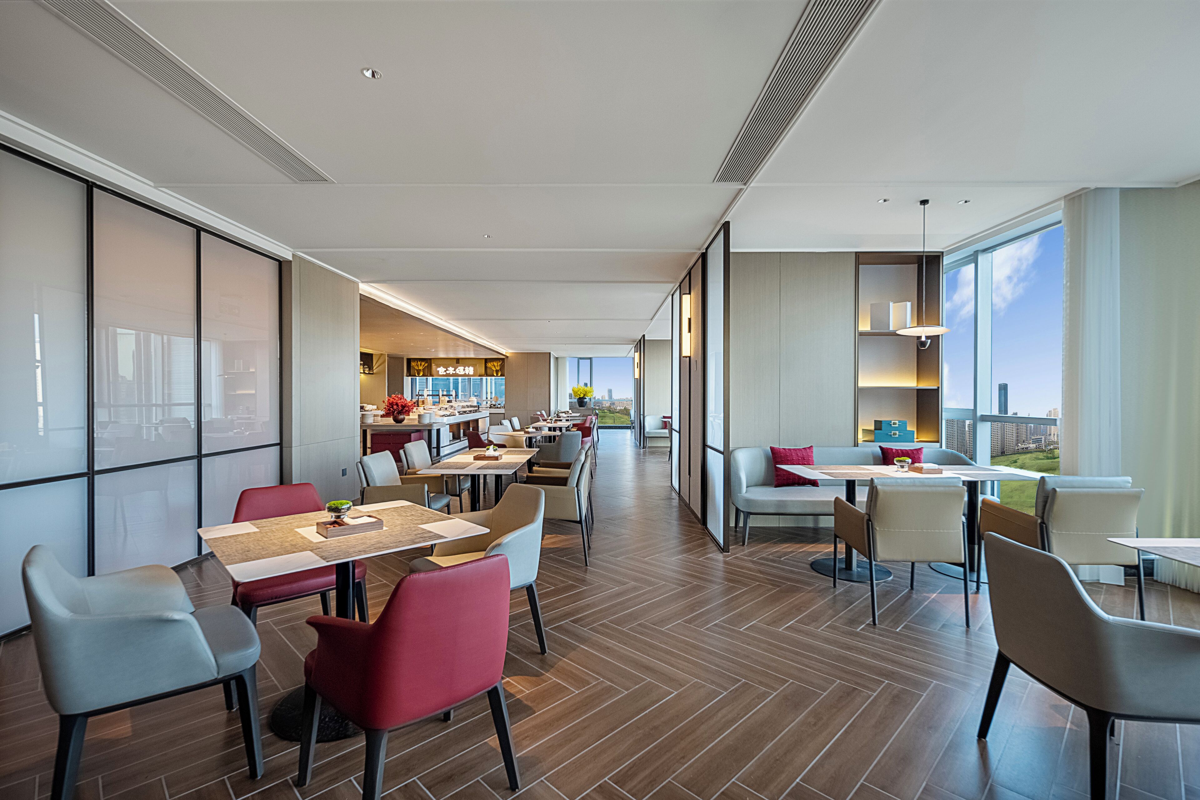 A modern hotel lobby with a long dining area, a kitchen in the background, and large windows offering a city view.