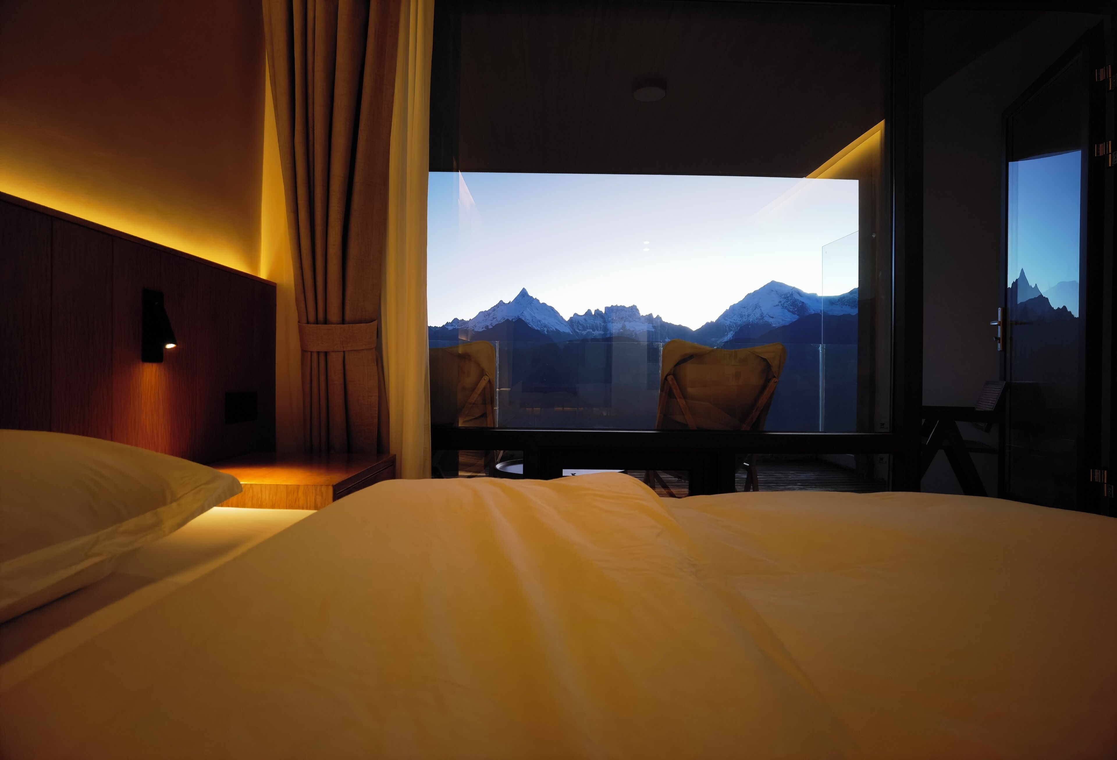 A hotel room with a bed, a desk, and a chair. There is a large window with a mountain view.