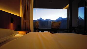 A hotel room with a bed, a desk, and a chair. There is a large window with a mountain view.