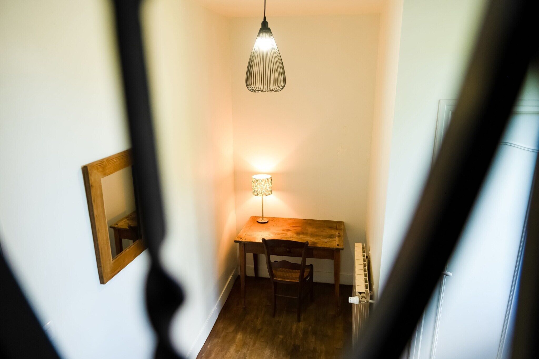 Un couloir étroit avec un bureau et une chaise en bois, un luminaire suspendu et un miroir au mur.