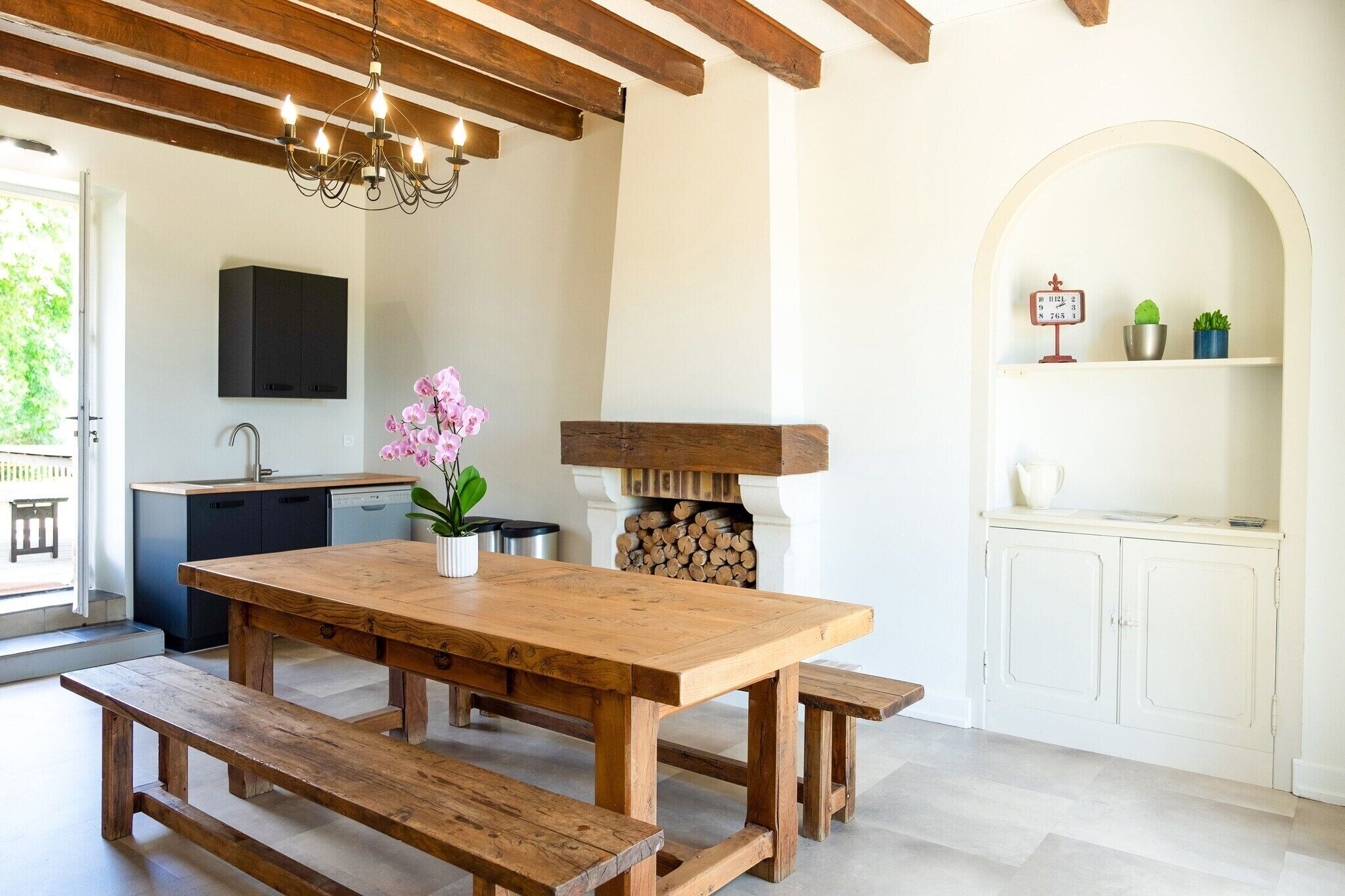 Une table à manger en bois avec des bancs, un lustre, une cheminée et un coin cuisine équipé de placards et d’un évier.