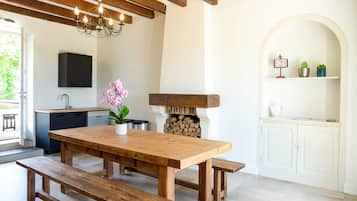 Une table à manger en bois avec des bancs, un lustre, une cheminée et un coin cuisine équipé de placards et d’un évier.