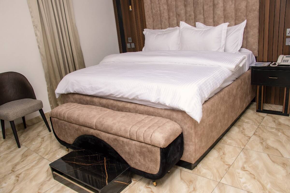 A hotel room with a large bed, a bedside table with a phone, a chair, and a bench.