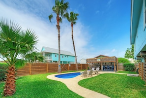 Um quintal com piscina, palmeiras, deck de madeira e uma casa com telhado azul.