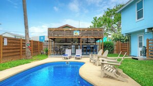 A swimming pool with lounge chairs, a covered patio, and a building with a sign. - Coastal Retreat w/ Pool,  BBQ & Sunset Views | Beers and Sunshine by AvantStay (Port Aransas)