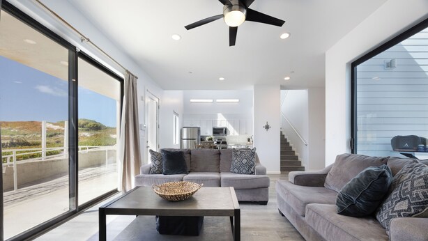 A modern living room with a large window, a ceiling fan, and a kitchen in the background. - Oceanfront 2BR + Rooftop Deck, & Beach Access | Strand Way B (Oceano)