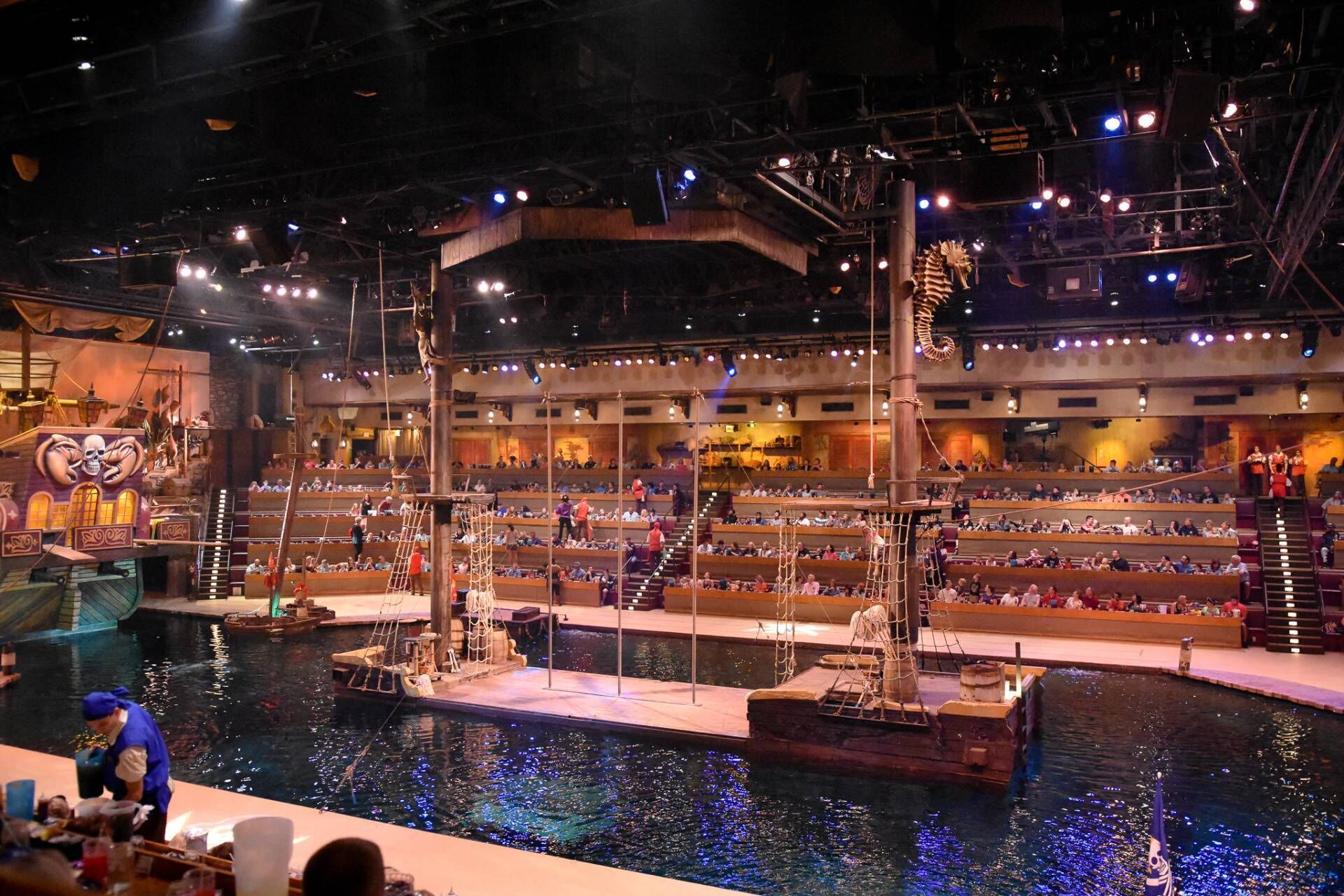 A large indoor venue with multiple levels of seating, a central stage, and a water feature with boats.