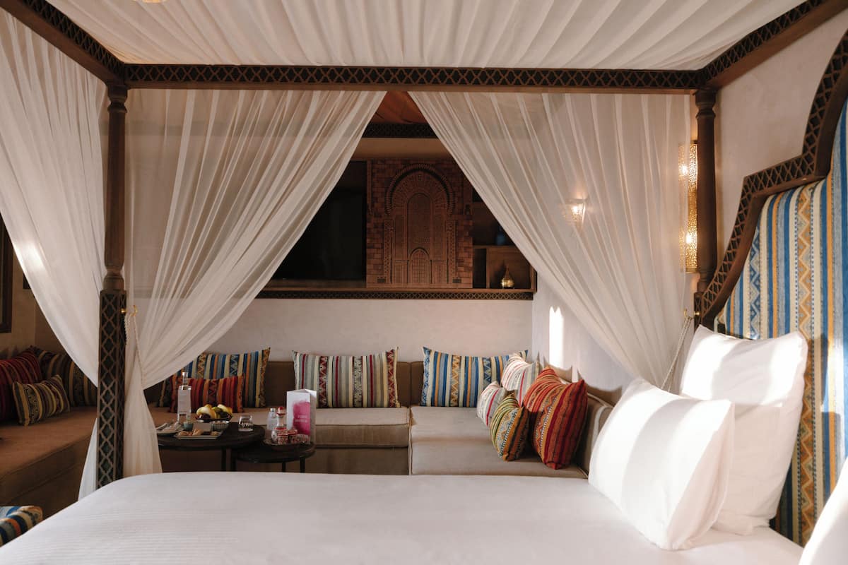 Ein Bett mit Baldachin, ein Sofa mit bunten Kissen und ein kleiner Tisch mit Obstschale und Flasche.