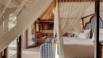 A four-poster bed with a canopy, a sofa, and a patterned armchair in a room with a wooden floor and a large window.