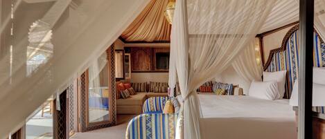 A four-poster bed with a canopy, a sofa, and a patterned armchair in a room with a wooden floor and a large window.