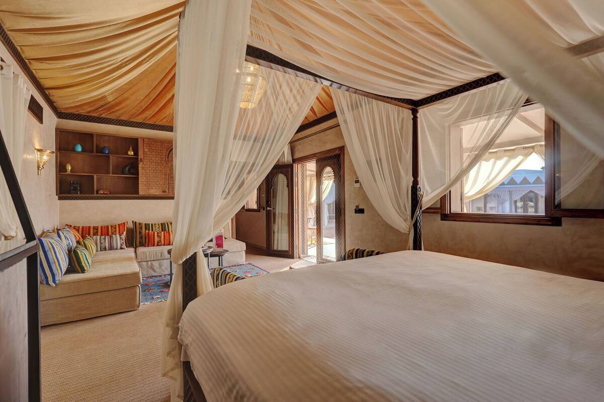 Ein Schlafzimmer mit Baldachinbett, einer Sitzecke mit Kissen und Blick ins Freie.