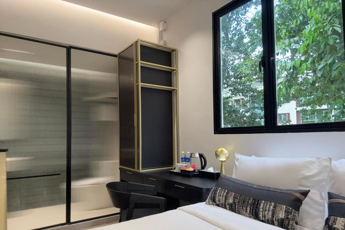 A modern hotel room with a bed, a desk, and a wardrobe. There is a window with a view of trees and buildings outside.