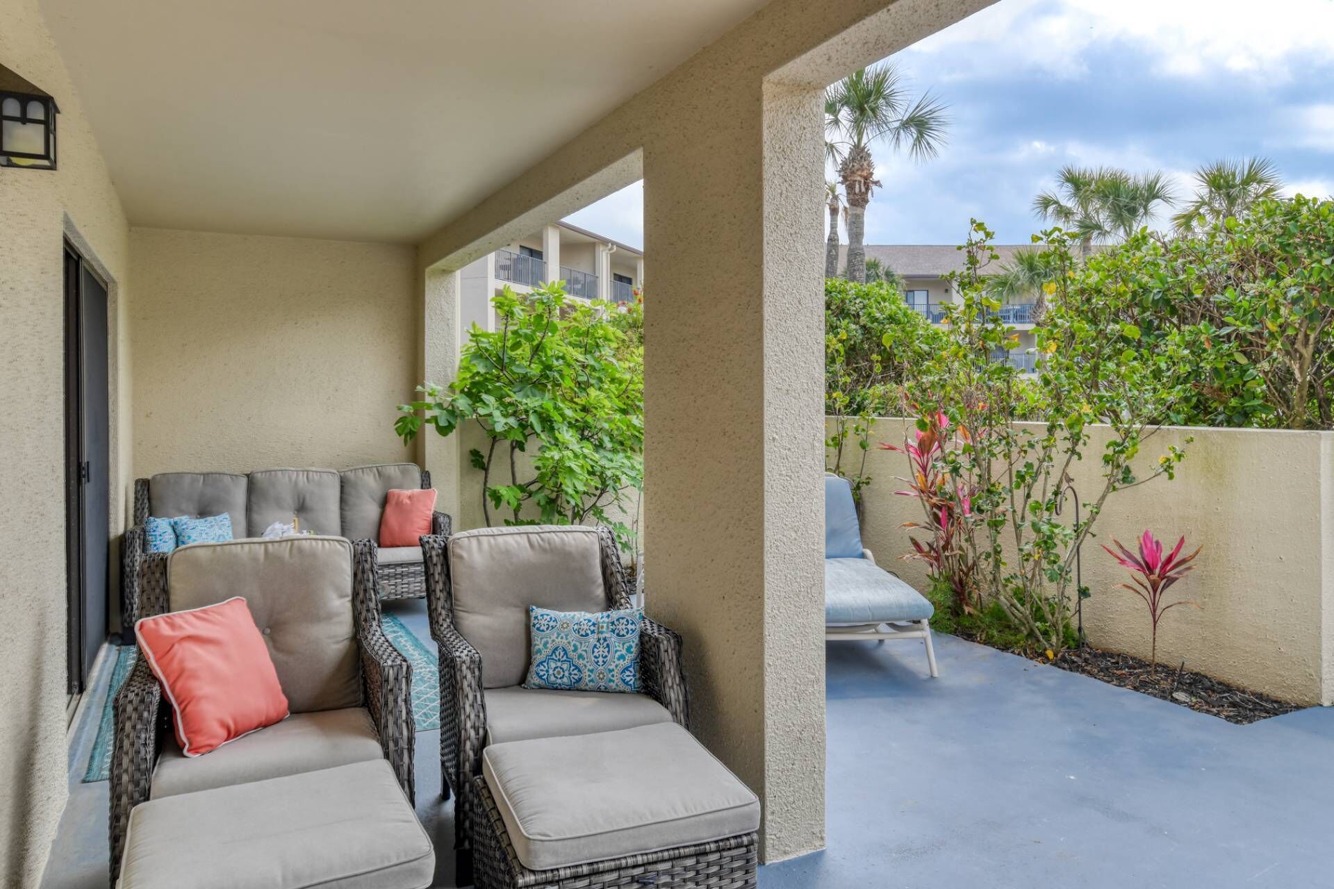 A covered outdoor seating area with wicker furniture and cushions, a concrete floor, and a view of a residential building and greenery.