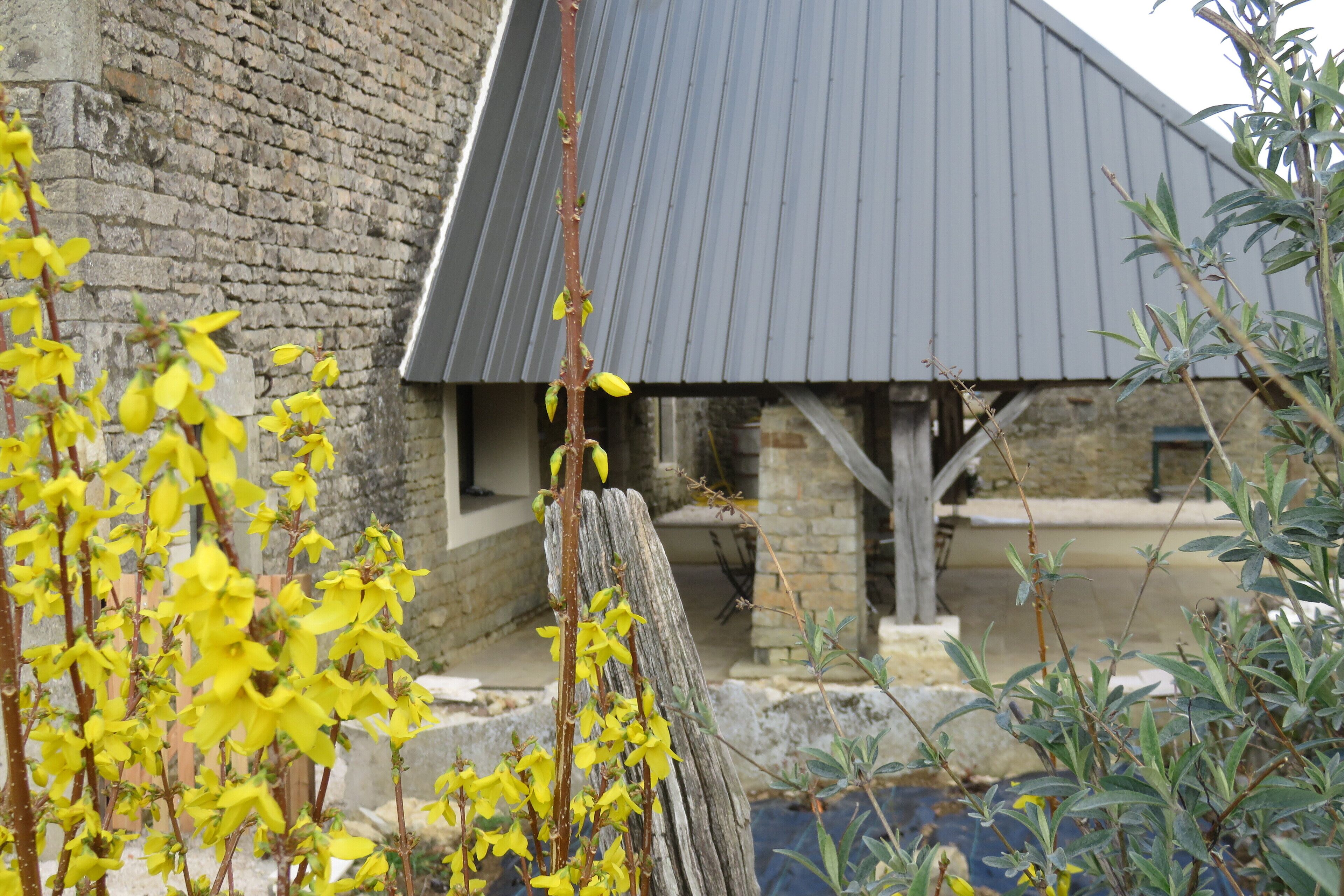 A rustic stone building with a metal roof, surrounded by greenery and yellow flowers.