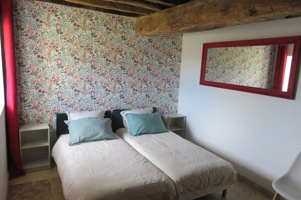 Un dormitorio con papel tapiz floral, una viga de madera en el techo, un espejo con marco rojo y dos mesitas de noche con almohadones.