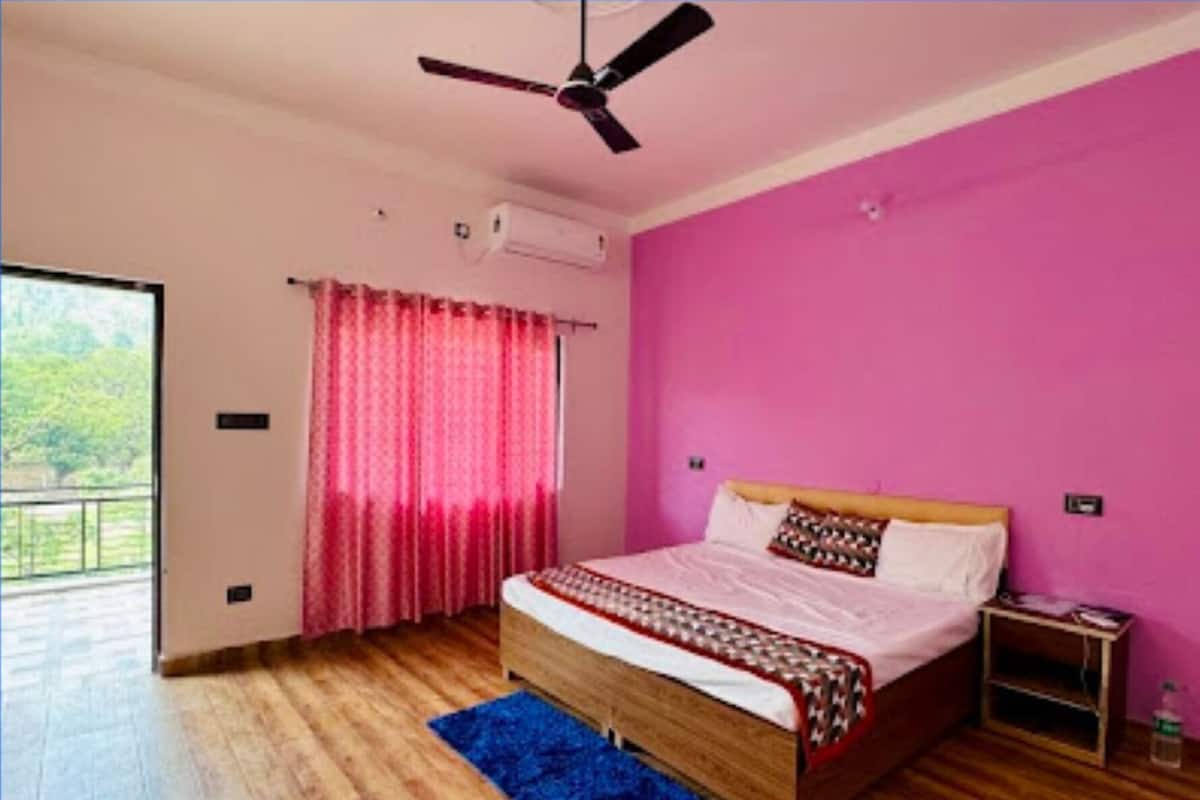 Une chambre avec un lit en bois, un ventilateur de plafond, un mur rose et une vue sur la verdure à l’extérieur.