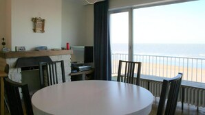 A room with a round table, chairs, a television, a fireplace, and a view of the beach.