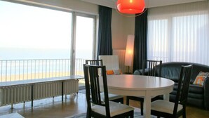 A room with a round table, chairs, a sofa, a lamp, and a view of the sea.