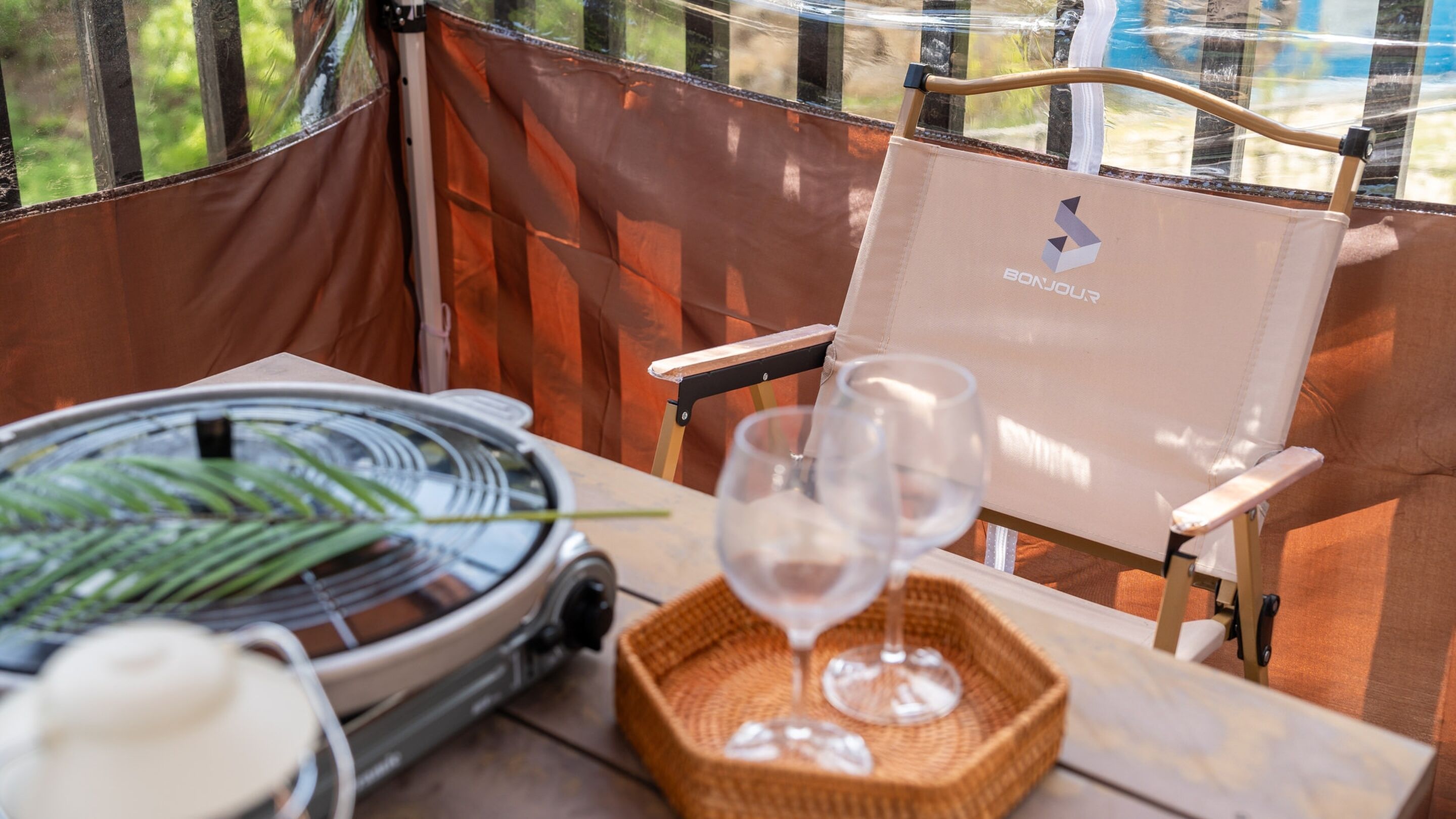 A dining setup with a grill, a basket, and wine glasses on a table inside a tent.