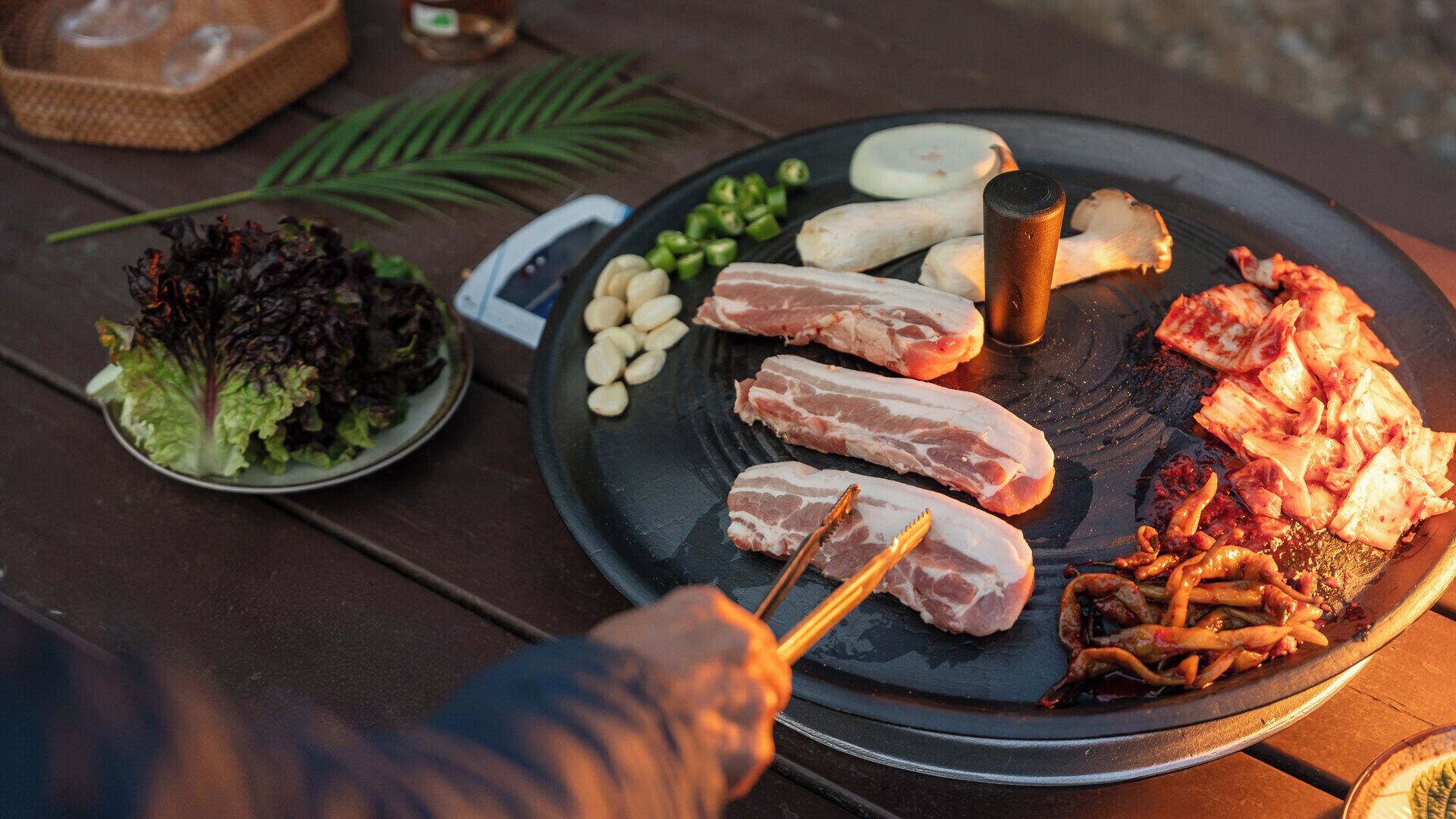 Een barbecue met verschillende vleessoorten en groenten, een saladekom en een fles saus op een houten tafel.
