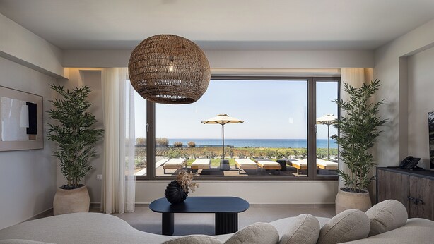 Een moderne woonkamer met een groot raam dat uitkijkt op een zwembad en de zee, een geweven hanglamp, en een pot met een plant.
