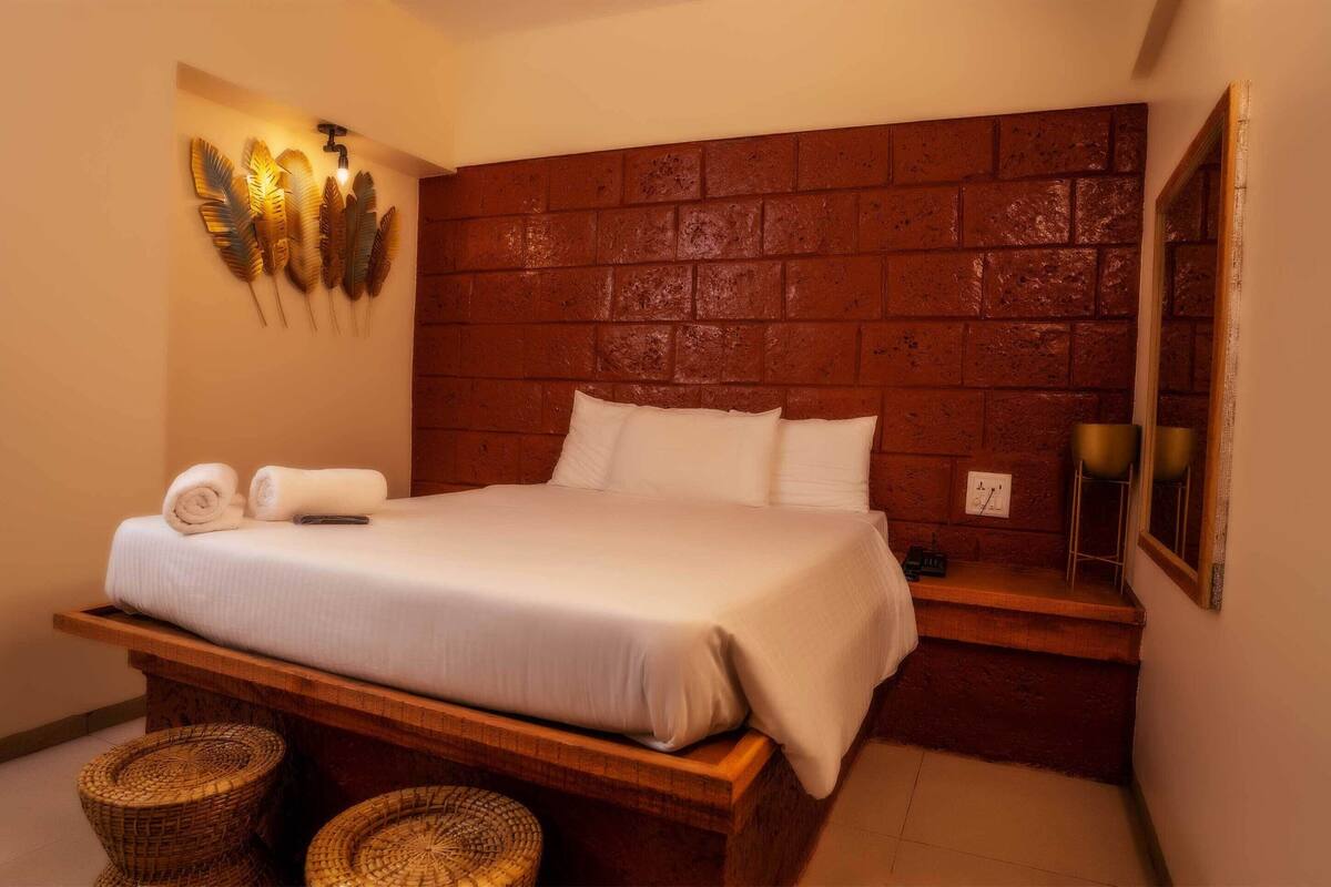 Quarto de hotel com cabeceira em padrão tijolo, cama com roupa de cama branca, dois cestos de vime e luminária fixada na parede.