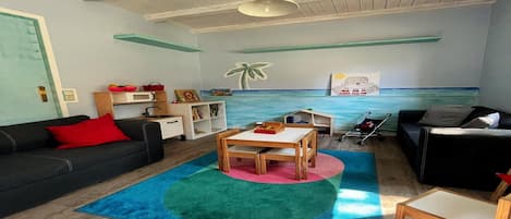 A living room with a wooden ceiling, a black sofa with a red pillow, a wooden table, and a colorful rug.