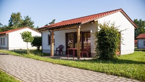 Un petit bâtiment d’un seul étage, doté d’un toit recouvert de tuiles rouges, d’un patio abrité avec deux chaises et une table, et d’un chemin pavé menant à l’entrée.