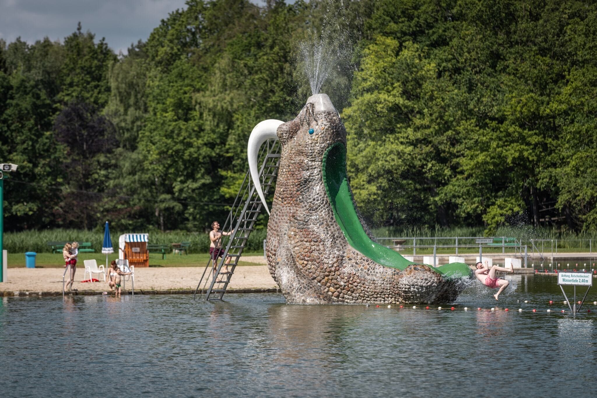 Un toboggan aquatique sur le thème des dinosaures, entouré d’une piscine et d’une zone sablonneuse avec des chaises et une échelle.