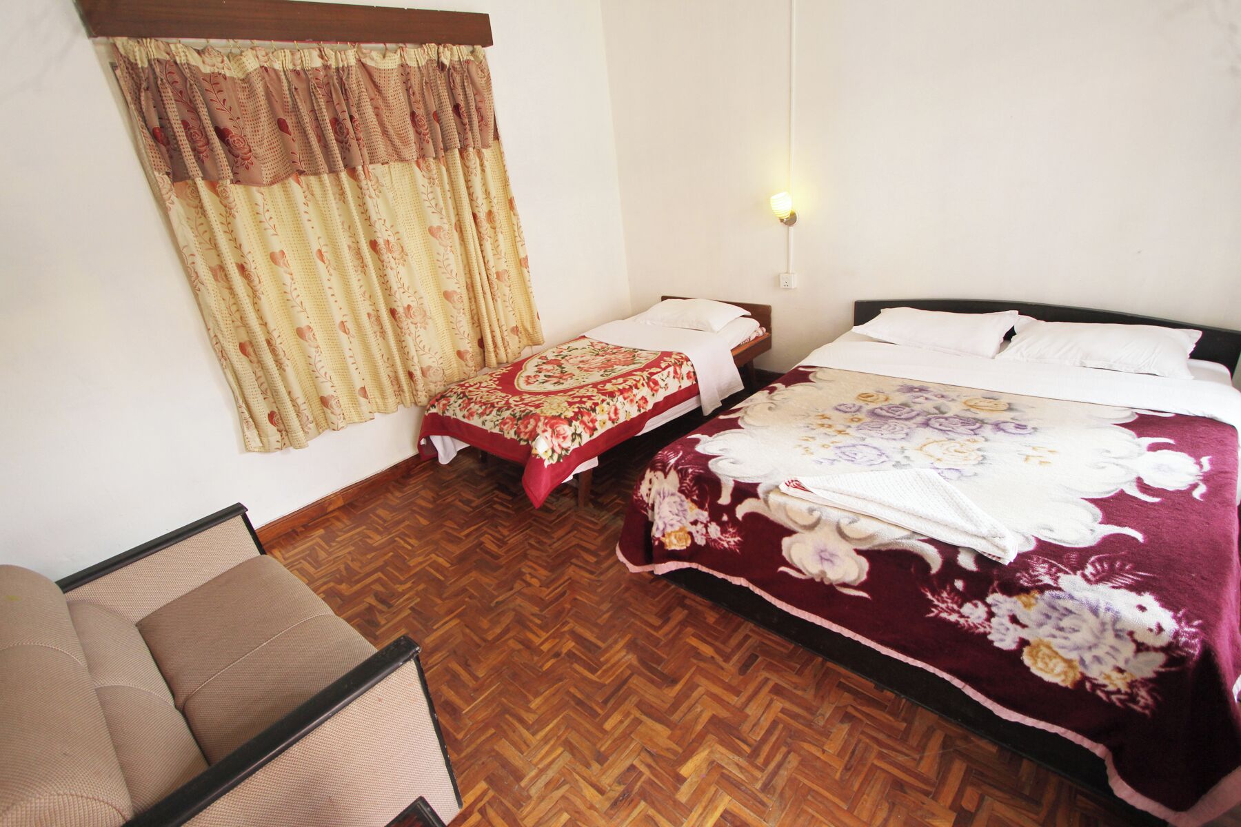 A hotel room with two single beds, a wooden herringbone floor, a window with patterned curtains, and a nightstand with a lamp.